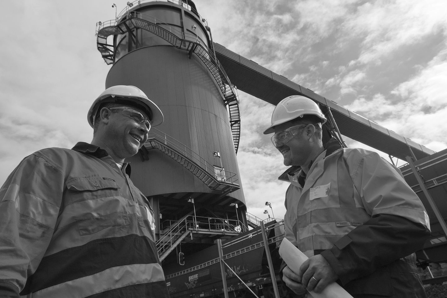 Two men in safety helmets and safety glasses stand on an industrial site