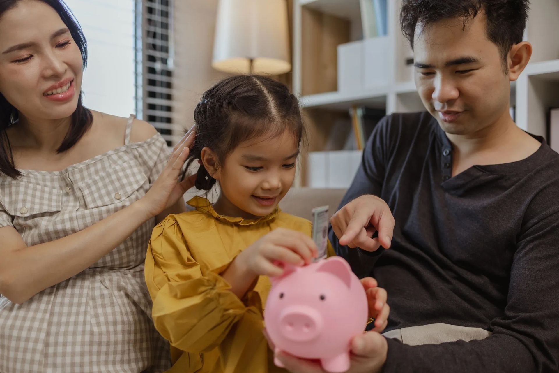 A happy family guiding their daughter as she puts money into a pink piggy bank at home. The parents smile while encouraging financial responsibility, symbolising eco-friendly habits, budgeting, and sustainable family living.