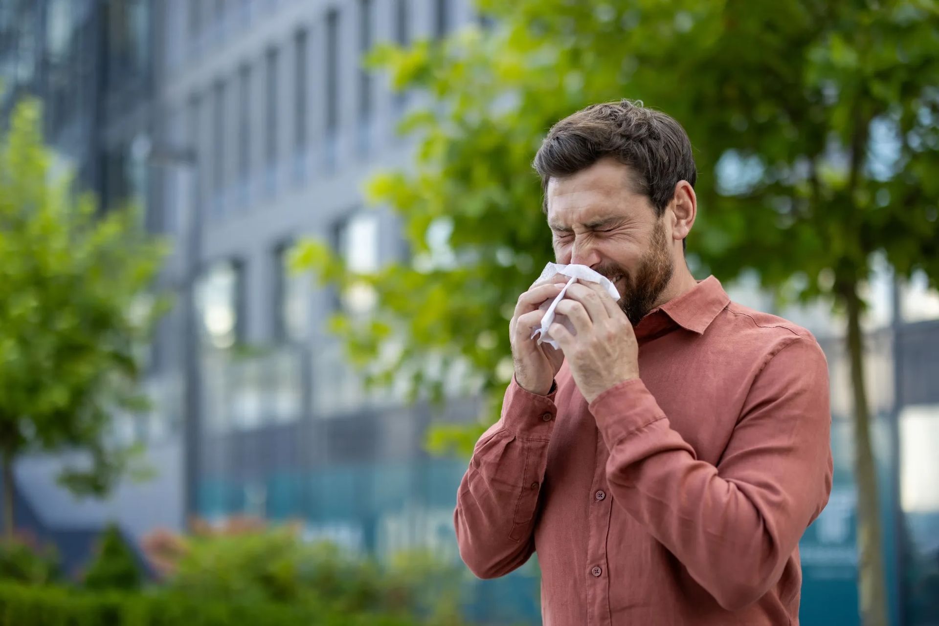 A man sneezing into a tissue while standing outside in an urban area with trees in the background. The image highlights seasonal hay fever, pollen allergies, and respiratory health in city environments.