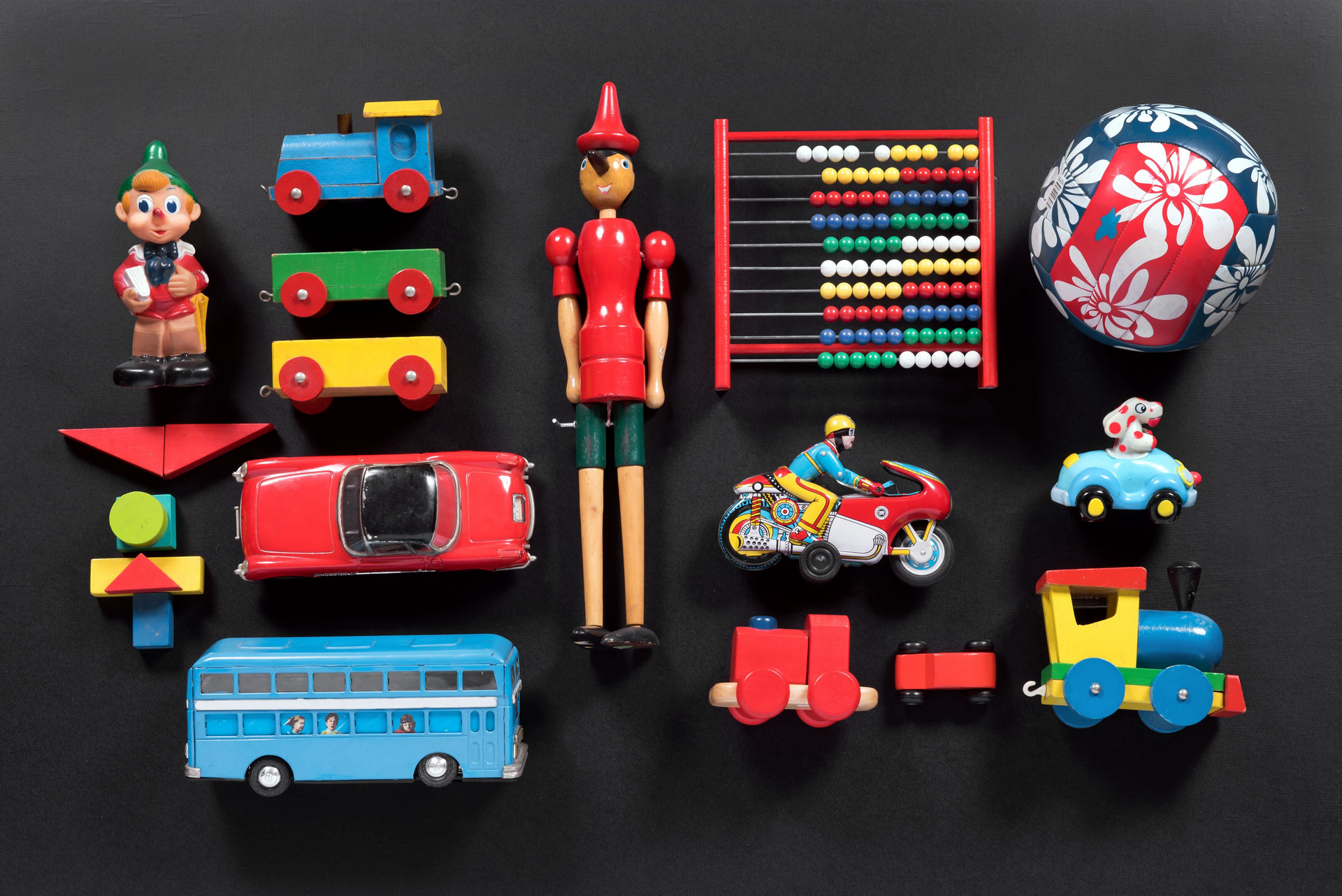 A colourful assortment of classic toys displayed on a black background, including a wooden Pinocchio doll, abacus, toy cars, trains, motorbike, ball, and a blue double-decker bus, evoking nostalgic childhood playtime.