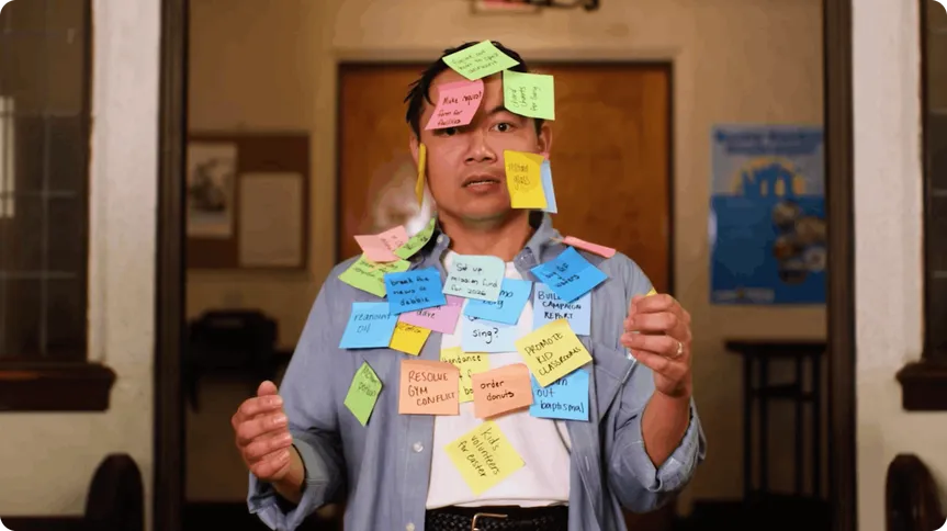 Person with a serious expression covered in colorful sticky notes with various tasks, standing in a room with a wooden door and a poster.