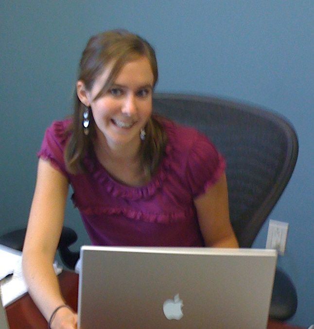 A smiling woman in a purple shirt sitting at a desk typing on a laptop.