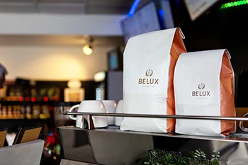 Two white Belux coffee bags with orange sides on a metal shelf in a cafe setting with cups and blurred background.