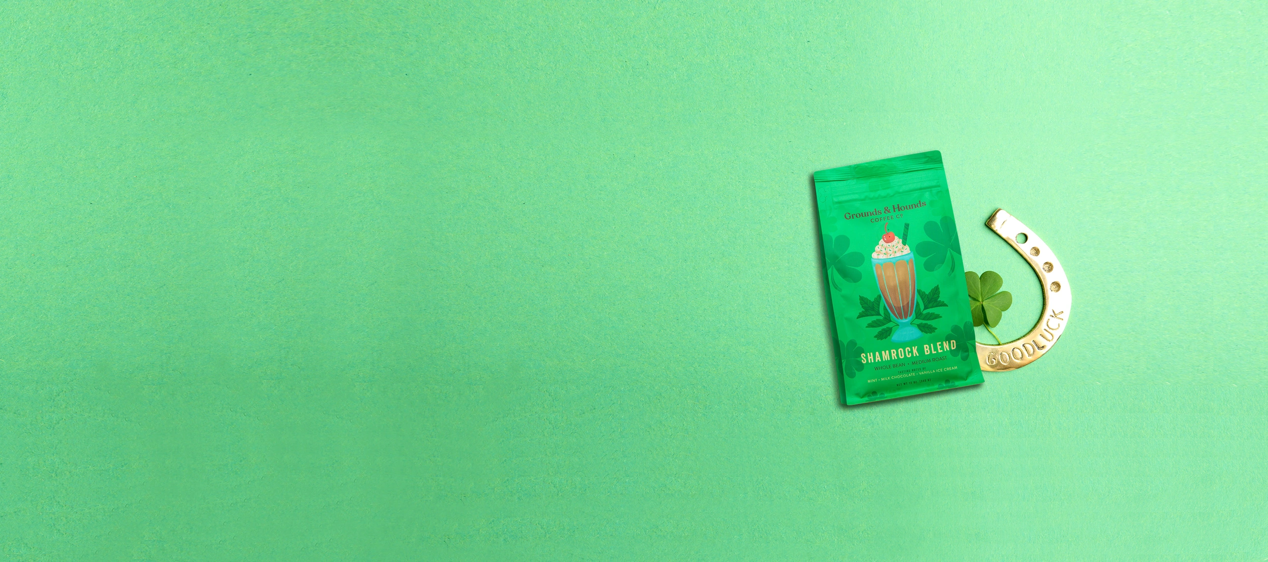 Green flat bottom bag labeled "Shamrock Blend" next to a clover and a gold horseshoe with "Good Luck" on mint green background.