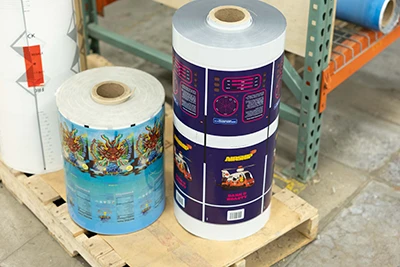 Two large rolls of colorful packaging rollstock standing on a wooden pallet in an industrial setting.