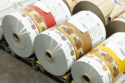 Large rolls of printed packaging material featuring colorful designs and labels, stacked horizontally on a pallet in an industrial setting.