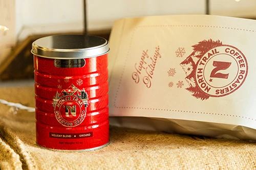 Red tin can of North Trail Coffee Roasters Holiday Blend with metallic effects next to a matching festive paper mailer bag on a rustic surface.