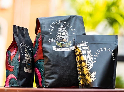 Three custom coffee bags from Seven Seas Roasting Co. featuring a ship and floral design, displayed outdoors.