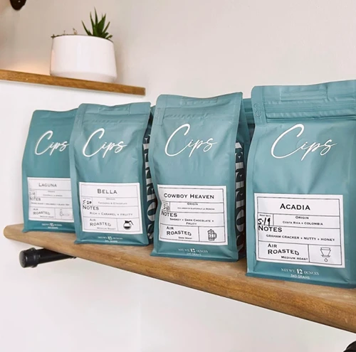Four teal custom flat bottom coffee bags labeled "Cips" on a wooden shelf, with a small plant in the background.