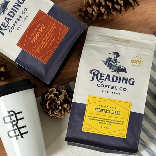Two Custom-Printed Flat Bottom Bags of Reading Coffee Co. coffee, "Pumpkin Spice" and "Breakfast Blend," on a table with pine cones and a white travel mug.