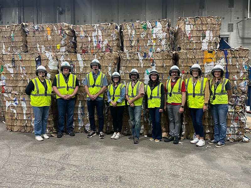 Group wearing safety gear stands in front of large stacks of compressed recyclables in a warehouse setting.