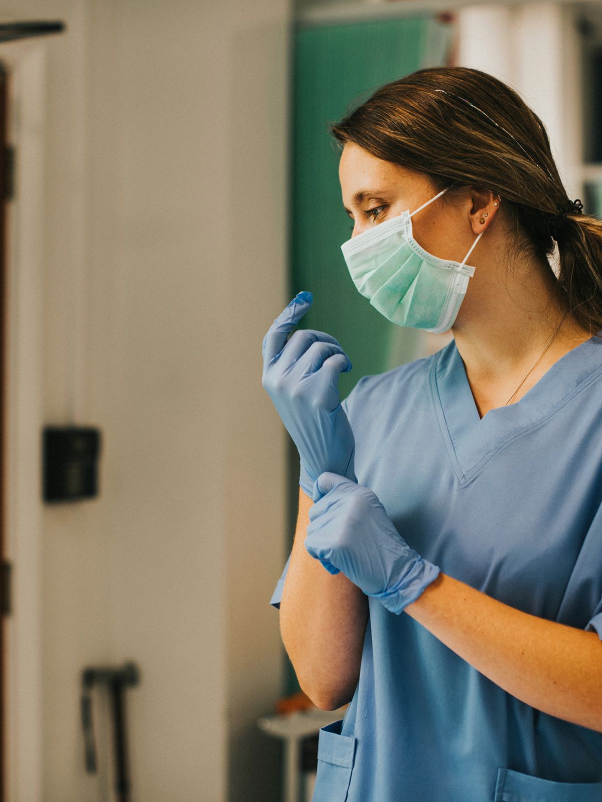 An independent nurse wearing blue scrubs and a surgical mask looks over her right shoulder as she pulls a glove onto her right hand