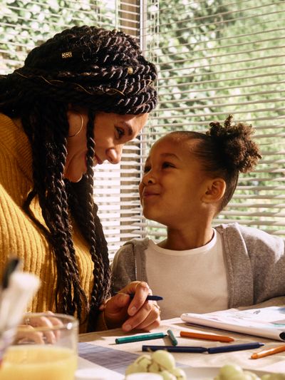 A female nurse and her young daughter look affectionately at each other while drawing pictures together at their kitchen table on a sunny morning