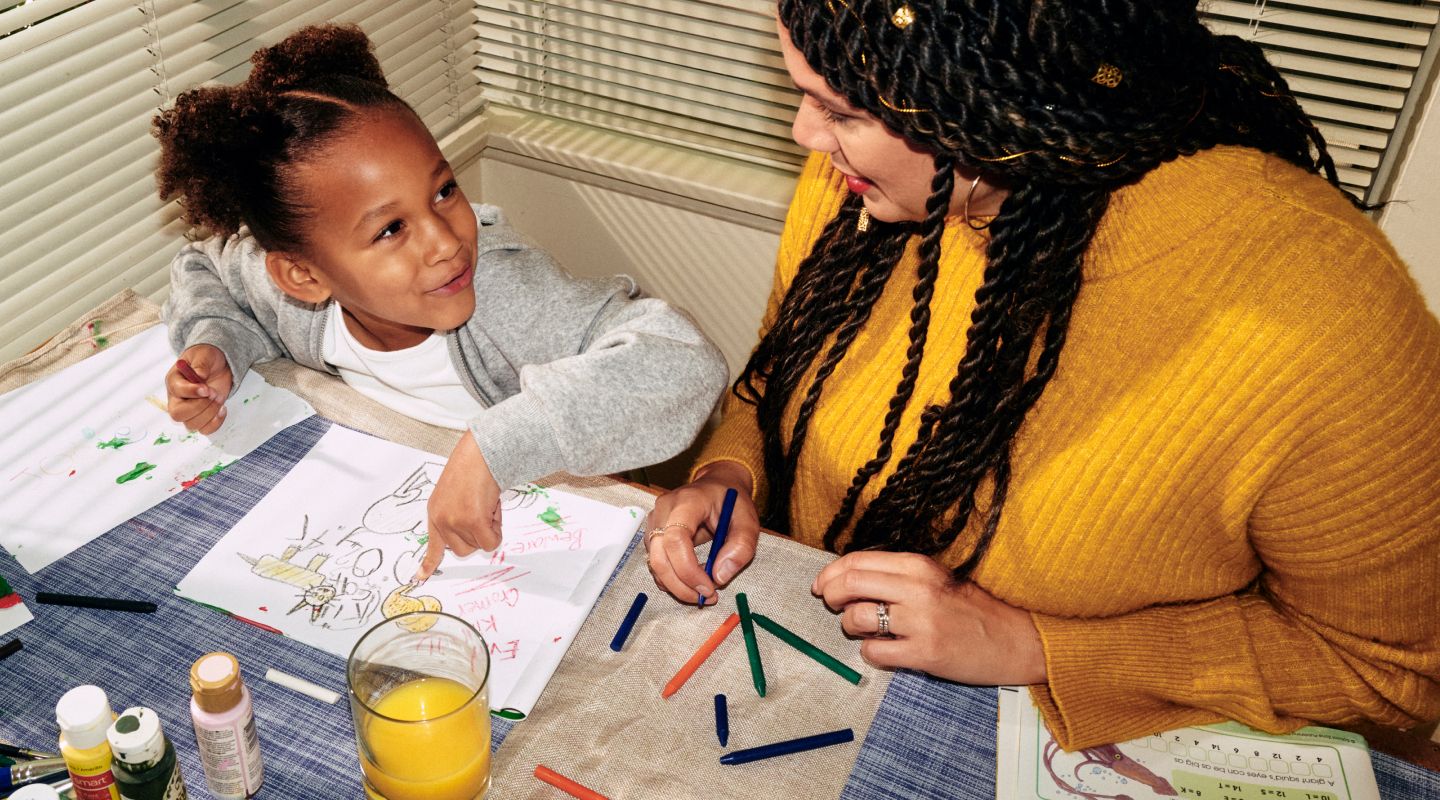 A female nurse and her young daughter smiling at each other. The daughter is pointing at a drawing they are making together at their kitchen table on a sunny morning