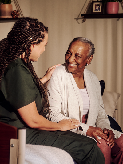 An independent professional wearing dark green scrubs places her hand on a smiling resident's shoulder.