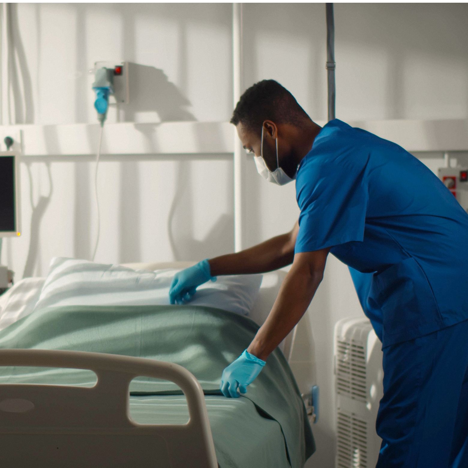 Masked independent nurse in blue scrubs wearing a mask and gloves making a hospital bed