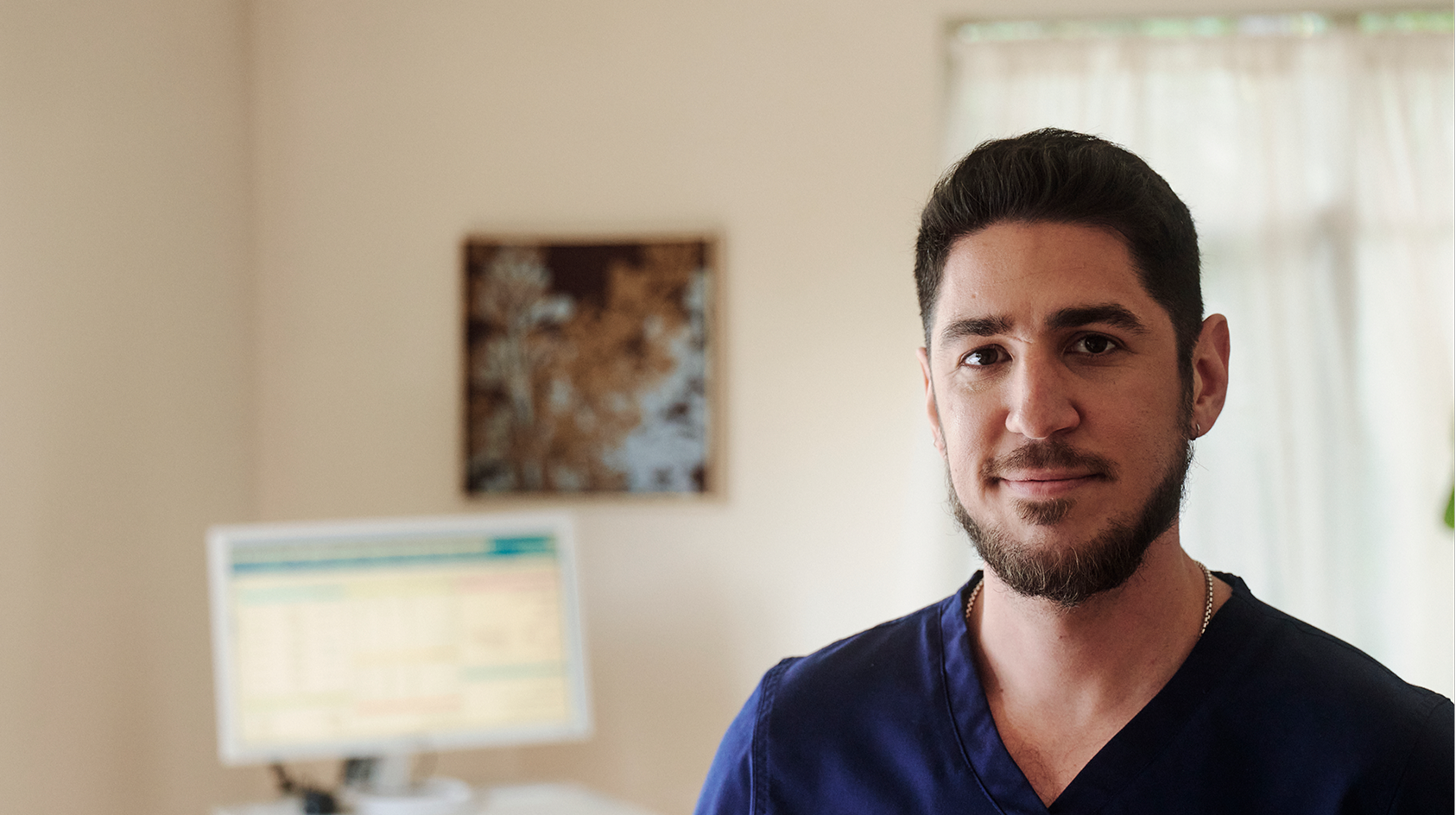 A headshot of a male nurse wearing blue scrubs with a computer behind him