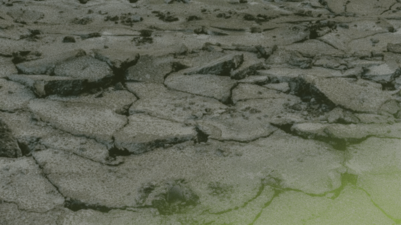 Cracked, dry earth with a rough texture, showing large, irregular pieces and a hint of green tint in the bottom right corner.