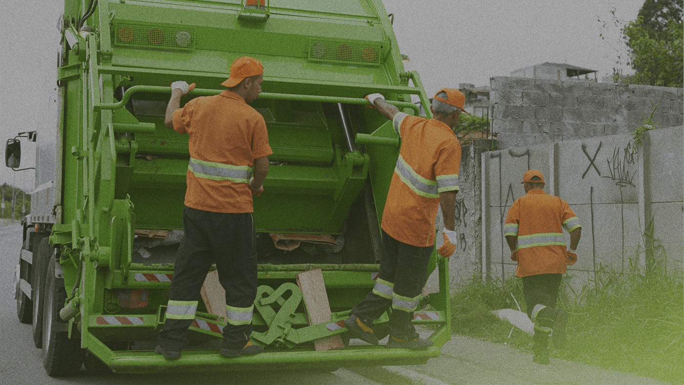 Three sanitation workers in orange uniforms operate a green garbage truck on a street, with one worker walking nearby.