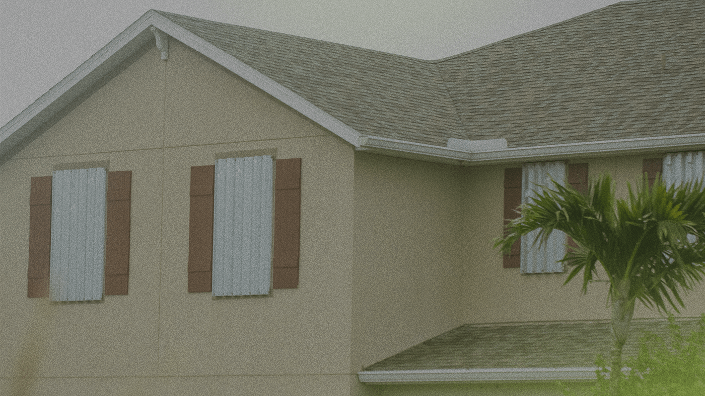A house with boarded-up windows and a palm tree in front, under a cloudy sky.