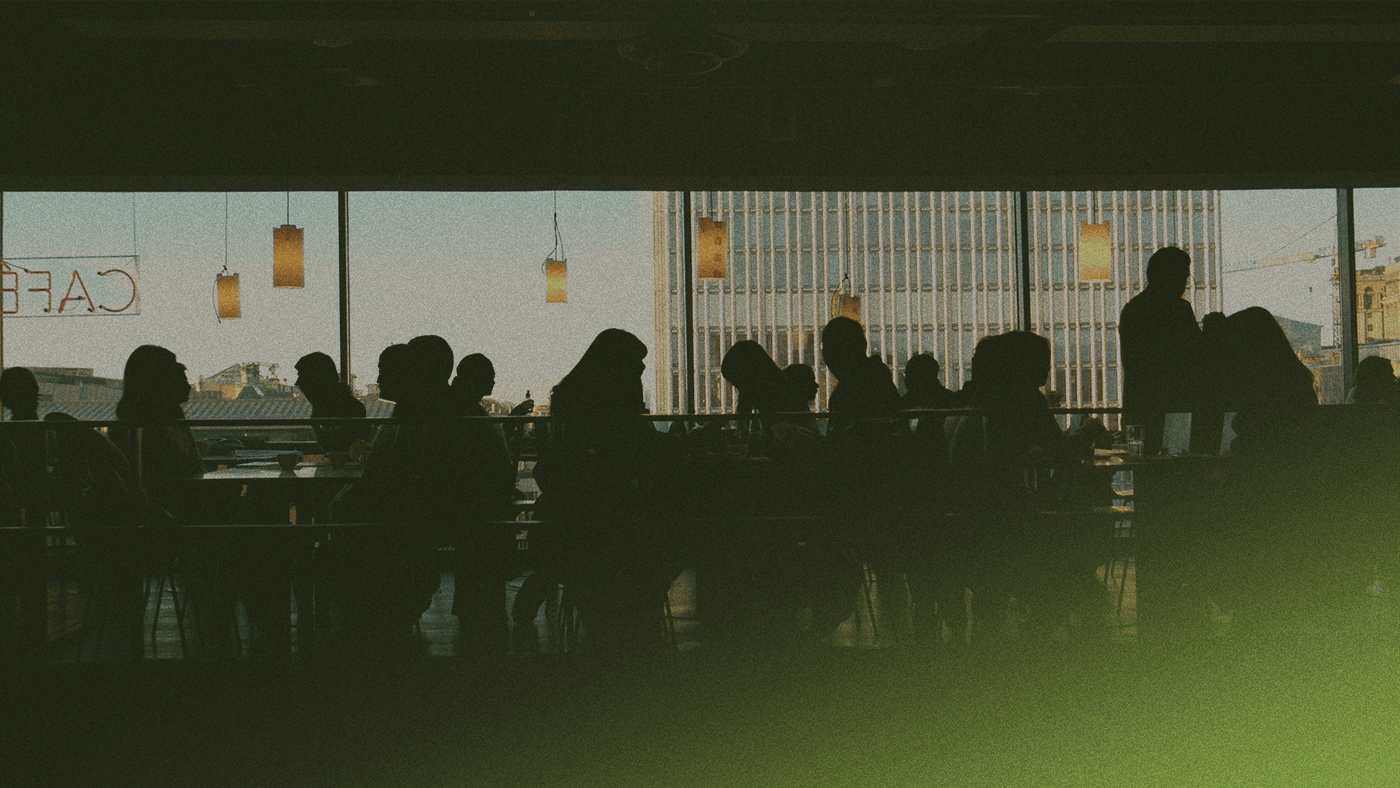 Silhouetted people sitting and walking in a cafe with large windows, soft lighting, and a view of buildings outside.