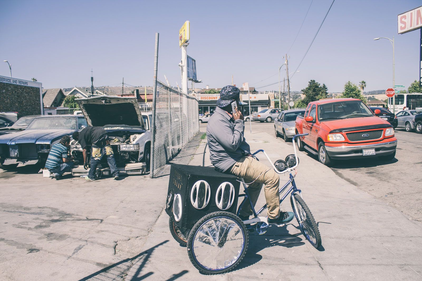 A parked cyclist is chatting on the phone. He bike is modified with three wheels and a black box of loud speakers.