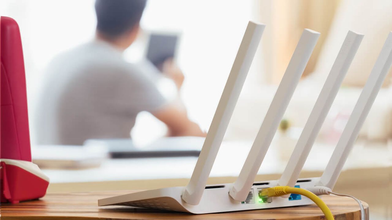 A white router is sitting on a desk while a man uses a tablet in the background