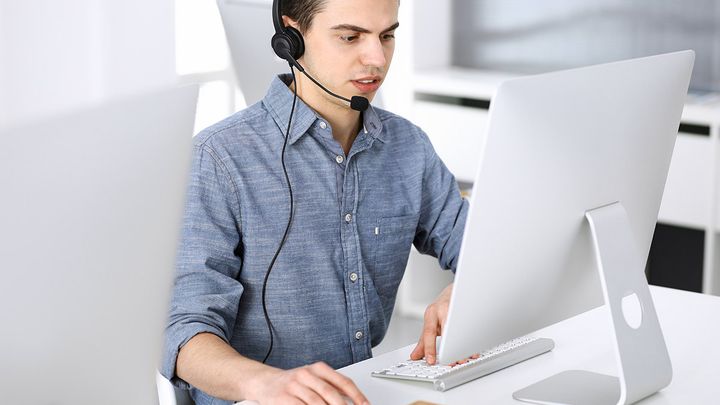 A man sitting at a desk looking at a computer screen and holding a computer mouse. He is wearing a headset with a microphone and appears to be talking into the microphone.