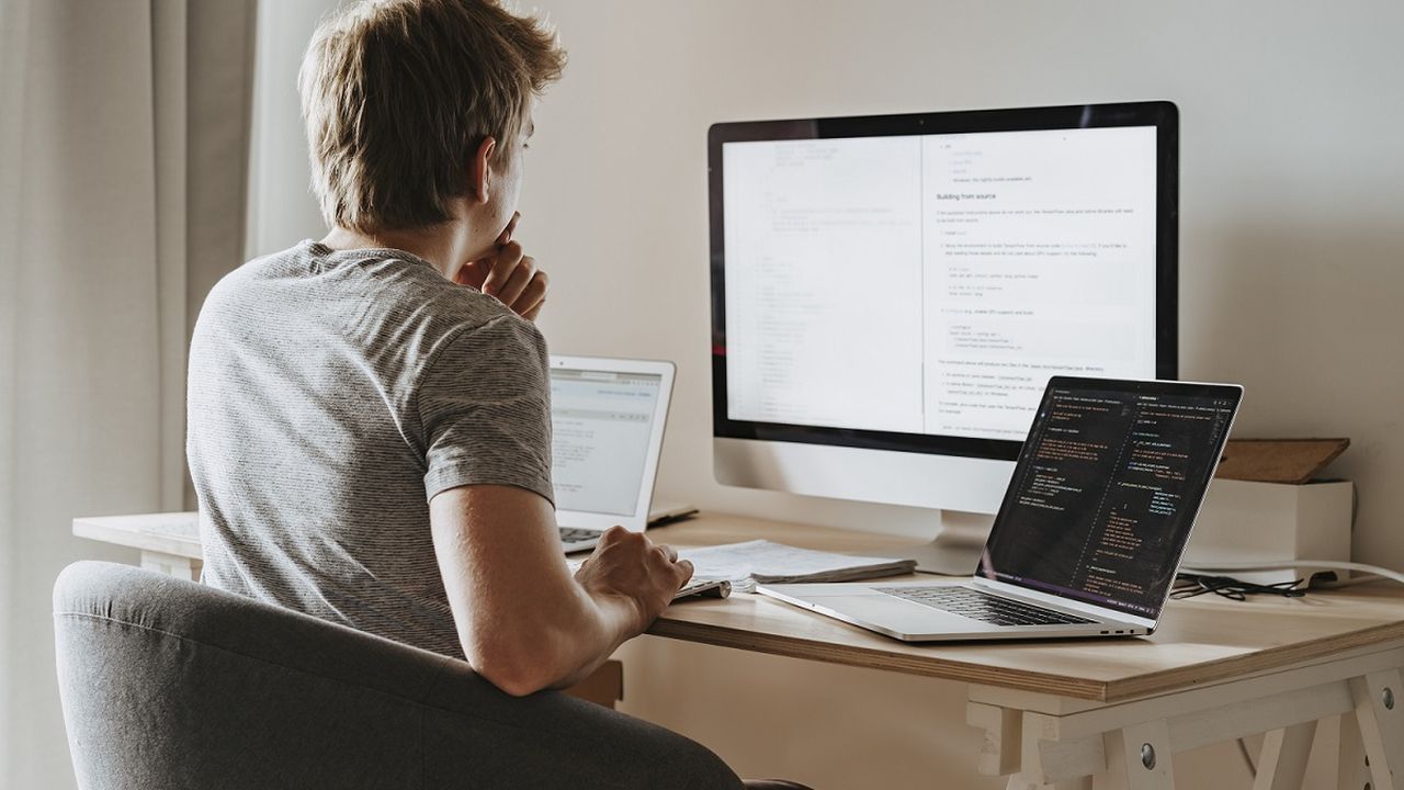 A person sitting at a desk thinking, while looking at a laptop and computer screen,.