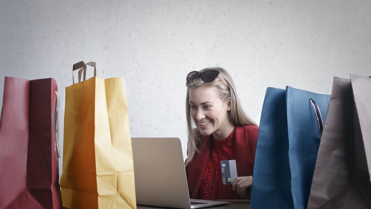 A woman sitting on a table smiling and looking at a laptop. She is holding a credit card in her hand. There are four shopping bags on the table, two on either side of the woman.