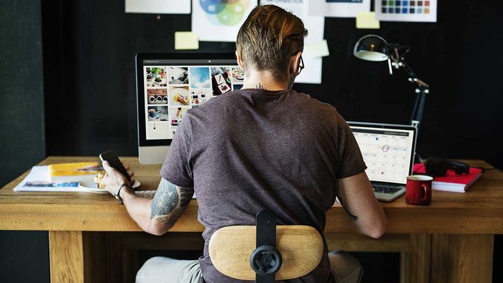 Stock photo of a person sitting at a desk using a computer, phone, and laptop