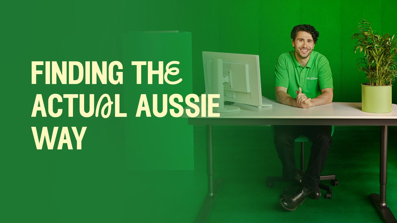 An image of a male sitting behind a desk in a green polo against the same coloured background. He has a computer to his left, and a plant to his right, with the left-hand side of the image containing the phrase "Finding the Actual Aussie Way".