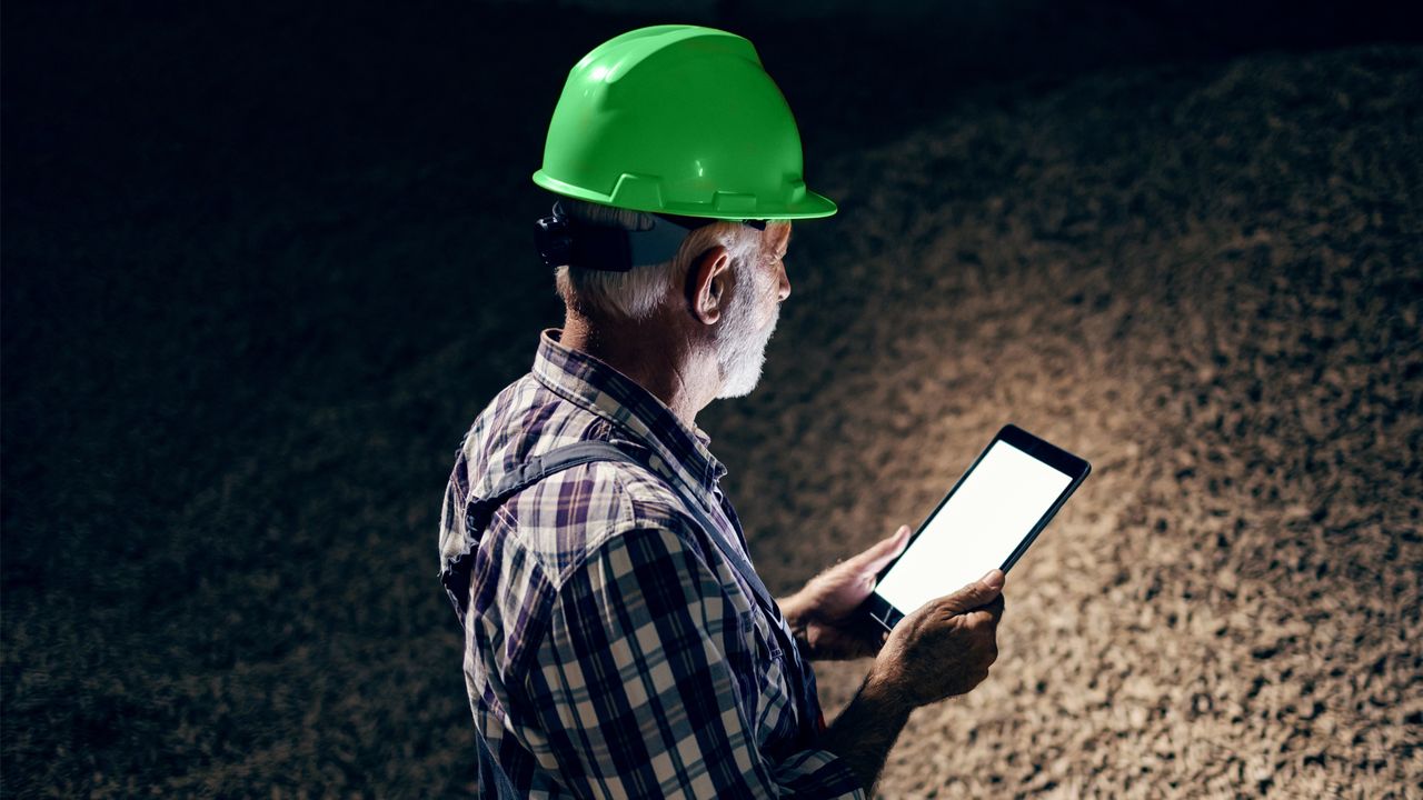 A man wearing a green hard hat standing outdoors surrounded by darkness, looking at a tablet, which provides the sole source of light in the image.