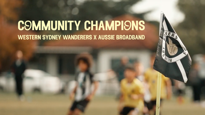 A soccer field with children playing in the background. A local club's corner flag is in the foreground with children players playing in the background on a field. Text reads "Community Champions Western Sydney Wanderers x Aussie Broadband"