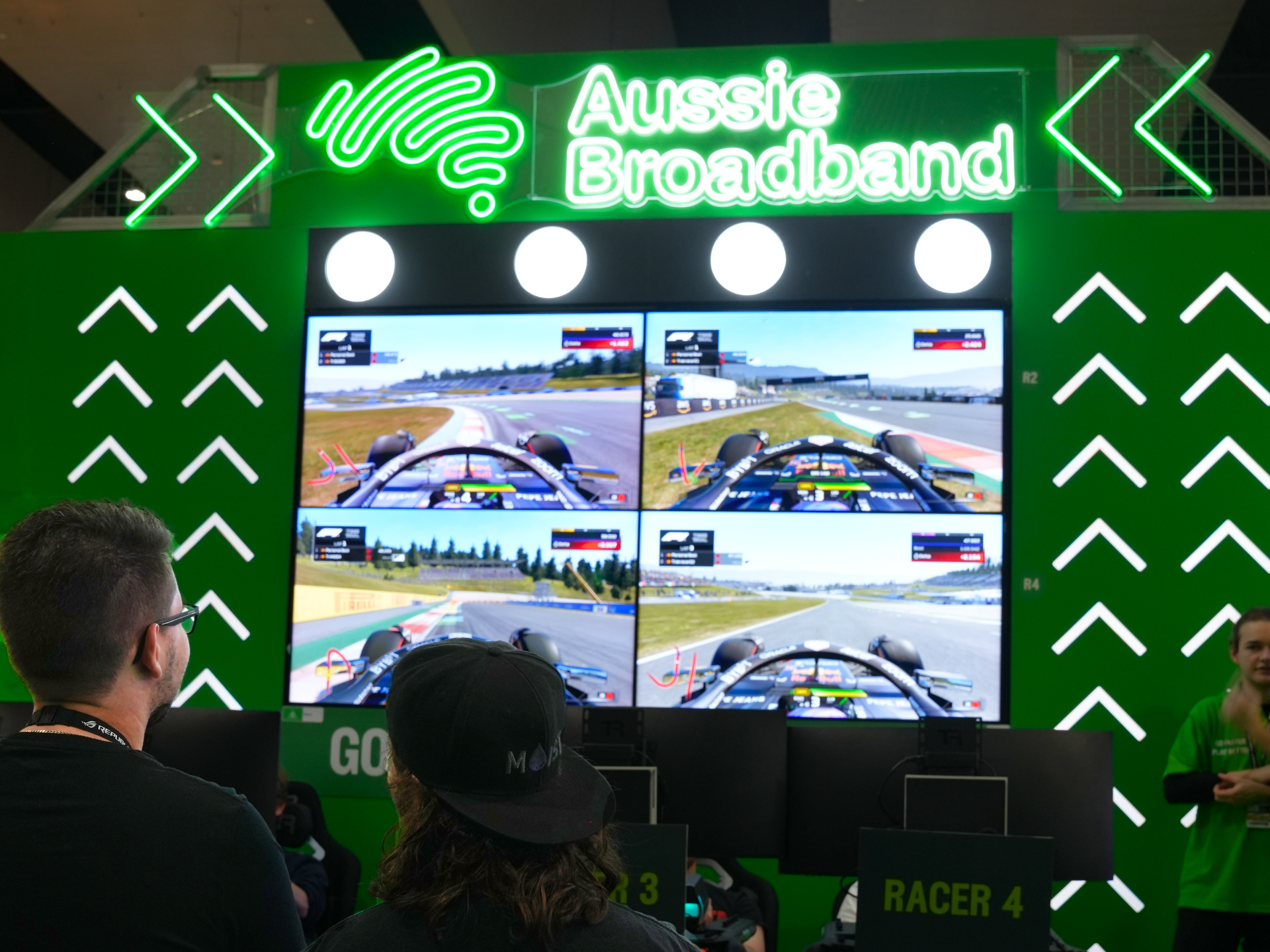 A photo of the Aussie Broadband expo booth wall at PAX with screens showing the Formula 1 game on them, with 2 people in the foreground looking at the screen.