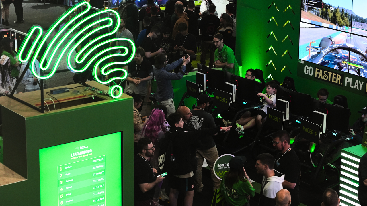 A photo of Aussie Broadband's PAX booth, with a neon squiggle logo sign and a screen leaderboard in the foreground, and a crowd of people and simulator setups in the background.