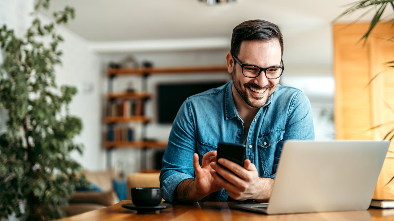 Photo of a person at a dining table, smiling while holding a smartphone and looking at a laptop in front of them