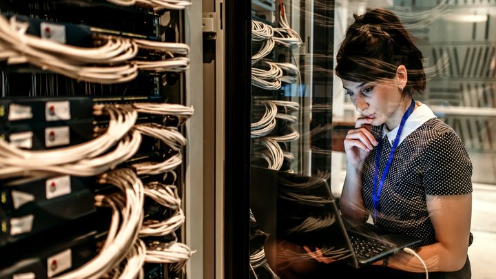 A woman looking at a laptop while standing in front of a network server