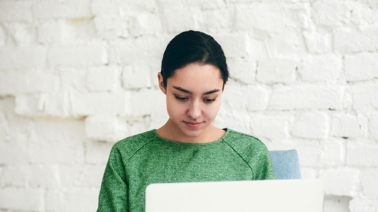 A photo of a female staring down at her laptop, in a green jumper and sitting down in front of a white wall.