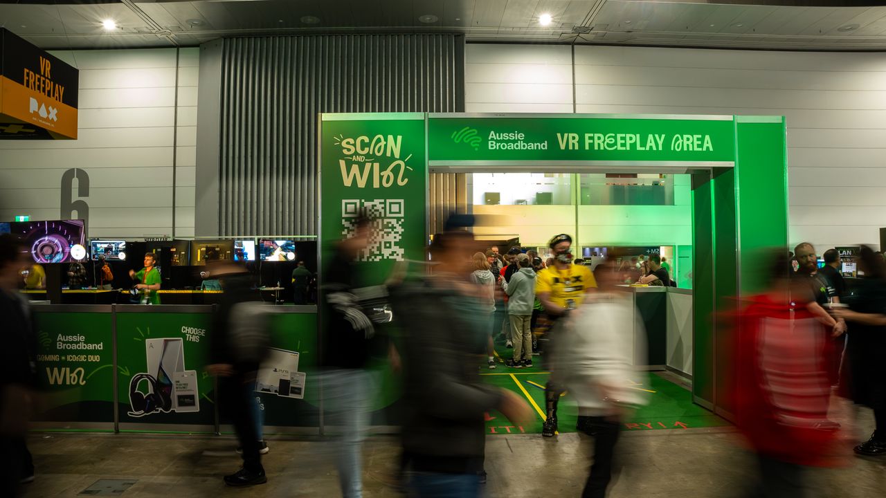 A photo of Aussie's PAX 2023 booth entrance, with a long exposure shot meaning the people walking in front are blurred. The archway is a green colour, with the text saying "VR Freeplay Area", "Scan to Win" (with a QR code underneath), and photos of gaming prize packs.