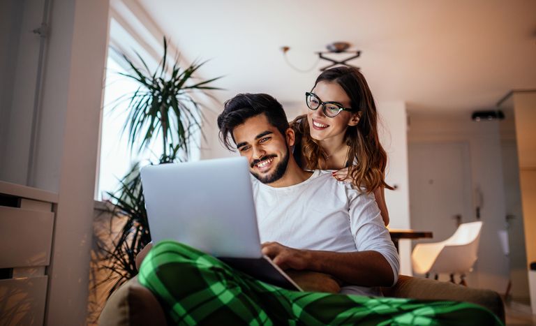 A couple looking at a laptop with the male in the foreground, lit by the laptop screen, and his female partner behind looking over his shoulder.