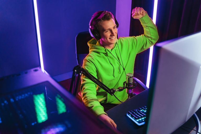 A male sitting in a dark, fluorescent-lit room with headphones, a microphone and a computer system in front of him. He is raising his fist and smiling, as if celebrating something he has seen on his computer.