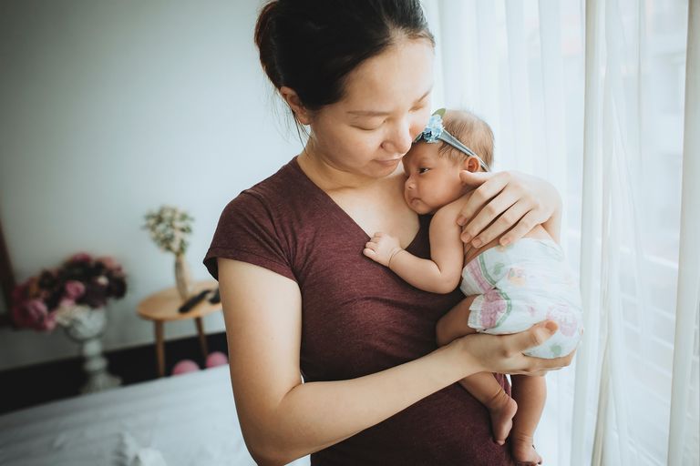 A mother with a red t-shirt on, holding her newborn infant in her hand. The infant has a blue headband and a nappy on, and they're seated in a room with a curtain and flowers in the background.