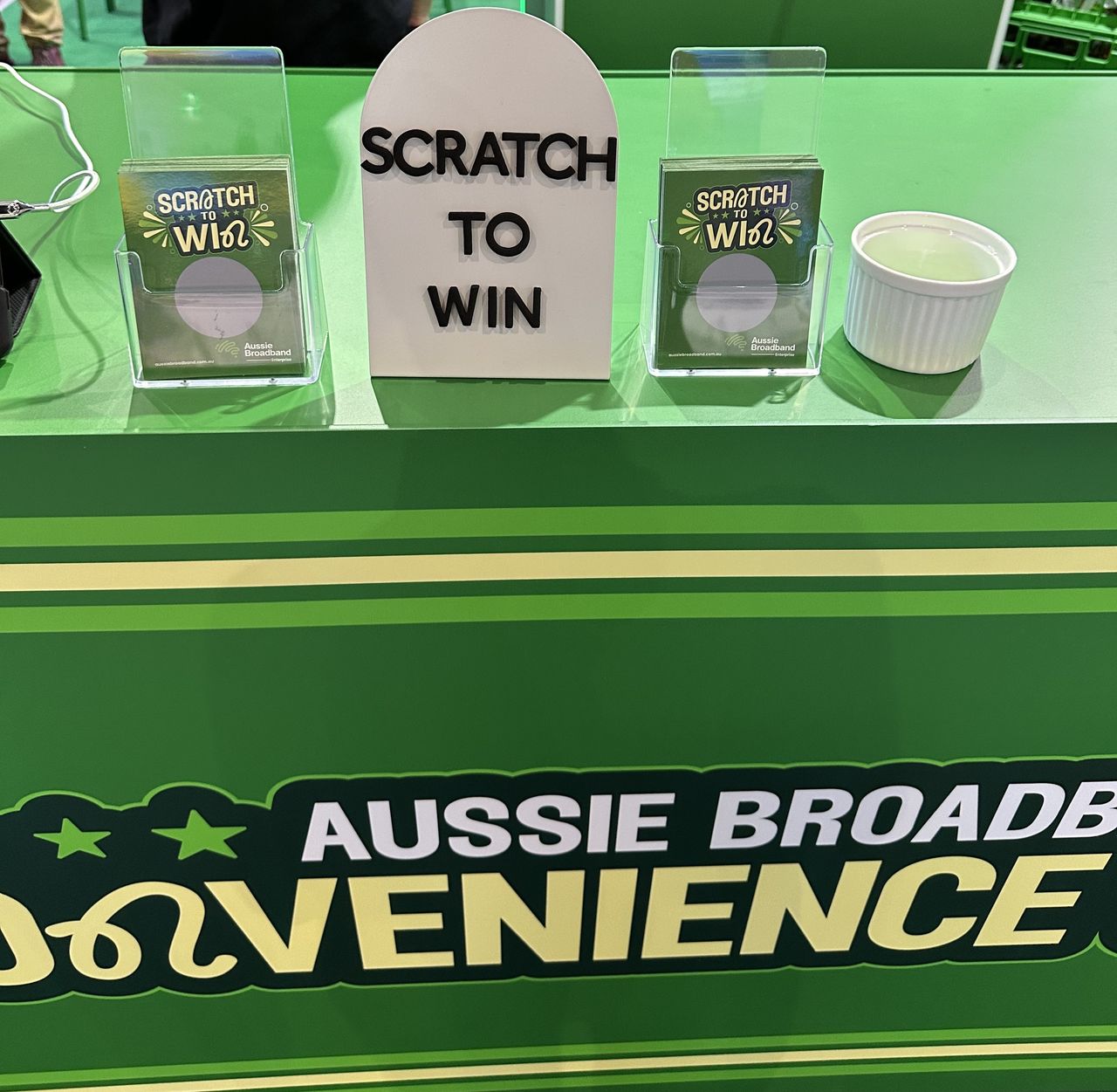A close up of Aussie Broadband's Cisco Live 2023 booth: A trade show booth resembling a convenience store. The front banner says "Aussie Broadband convenience store". It displays a "Scratch to win" competition, with a small sign saying "scratch to win". Either side of the sign are card holders containing scracties that say scratch to win and card holders that contain scratch cards. To the right is a tub that contains the scratchers contestants use to scratch the card and reveal their prize.