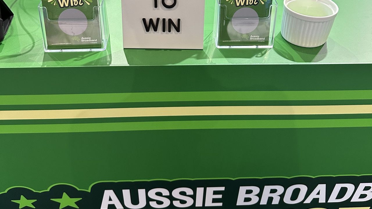 A close up of Aussie Broadband's Cisco Live 2023 booth: A trade show booth resembling a convenience store. The front banner says "Aussie Broadband convenience store". It displays a "Scratch to win" competition, with a small sign saying "scratch to win". Either side of the sign are card holders containing scracties that say scratch to win and card holders that contain scratch cards. To the right is a tub that contains the scratchers contestants use to scratch the card and reveal their prize.