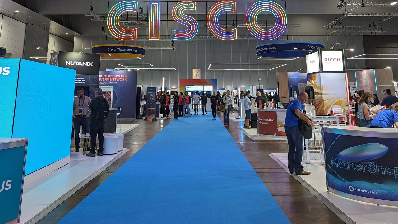 Cisco Live 2023: An image of a trade show, at the centre of the image is a blue carpet, and along either side of the carpet are various trade show booths. Above the booths is are illuminated letters that spell out "CISCO".