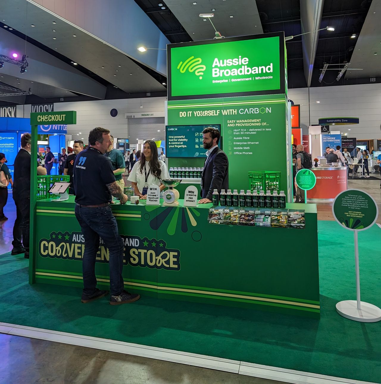 Aussie Broadband's Cisco Live 2023 booth: A trade show booth resembling a convenience store. The front banner says "Aussie Broadband convenience store" and there is a small, flag-like banner above that says "checkout". 2 people are behind the counter, and they are speaking to one person in front of the counter.