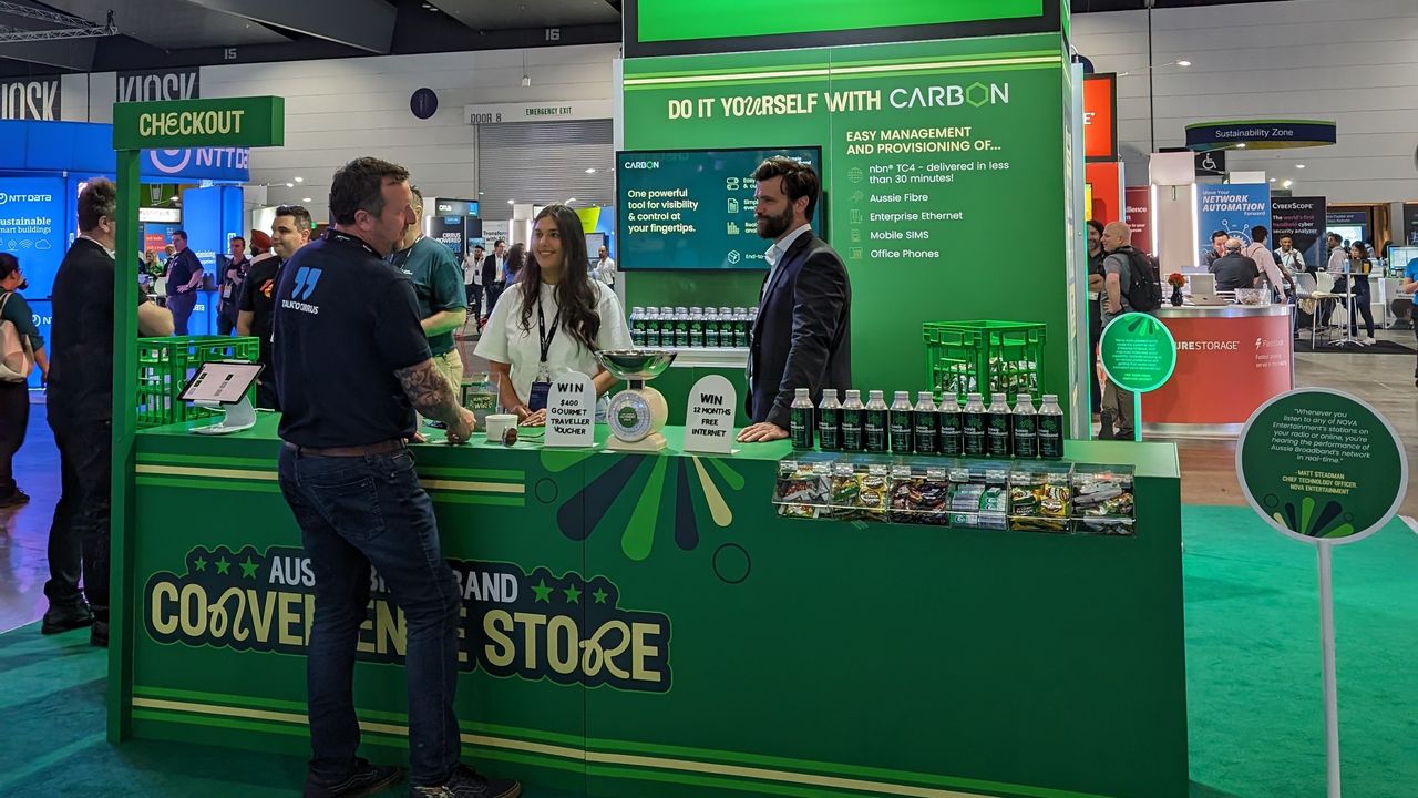 Aussie Broadband's Cisco Live 2023 booth: A trade show booth resembling a convenience store. The front banner says "Aussie Broadband convenience store" and there is a small, flag-like banner above that says "checkout". 2 people are behind the counter, and they are speaking to one person in front of the counter.