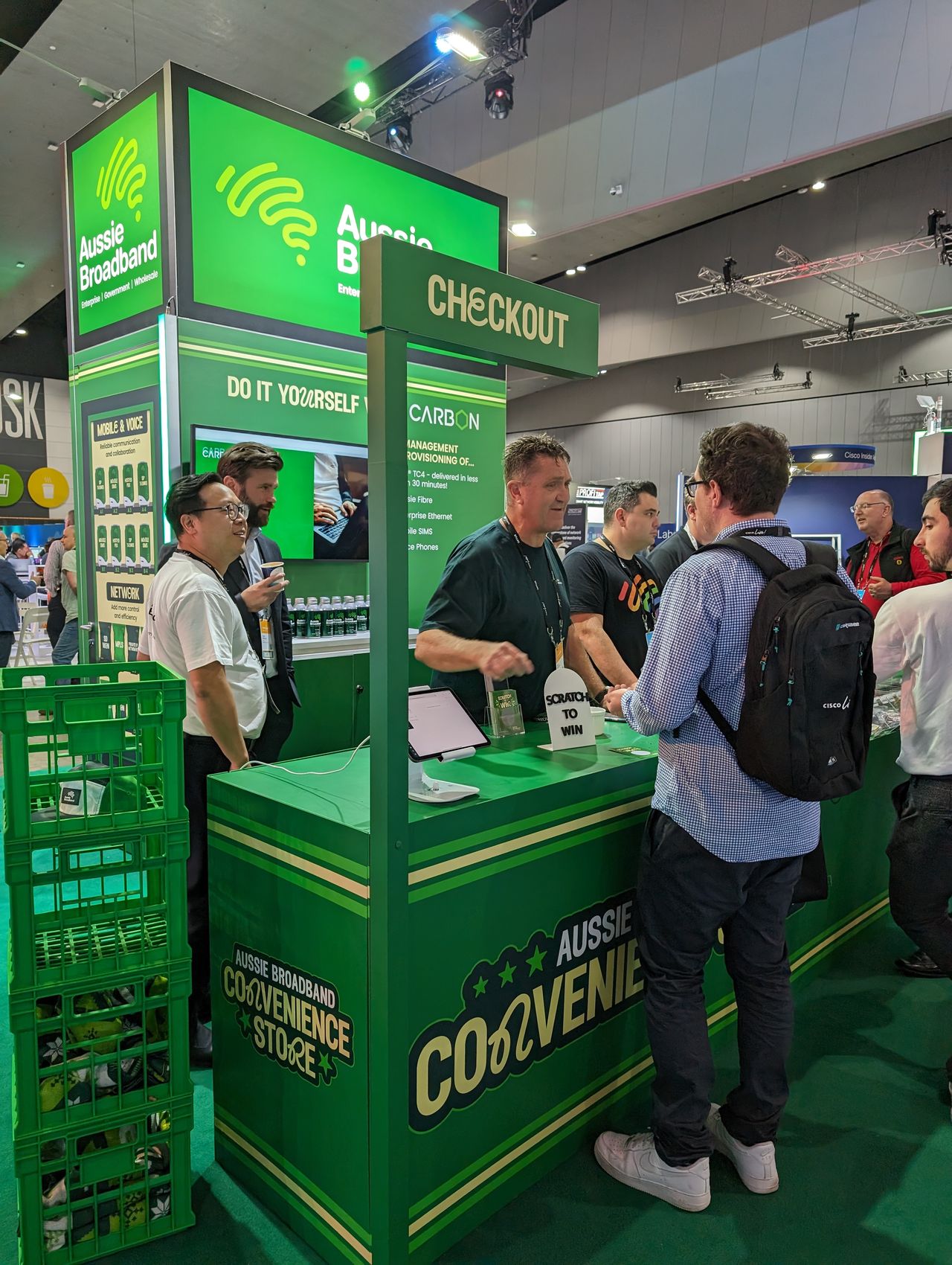 Aussie Broadband's Cisco Live 2023 booth: A trade show booth resembling a convenience store. The front banner says "Aussie Broadband convenience store" and there is a small, flag-like banner above that says "checkout". There are six Aussie Broadband staff members behind the counter, and three event attendees in front of the counter.