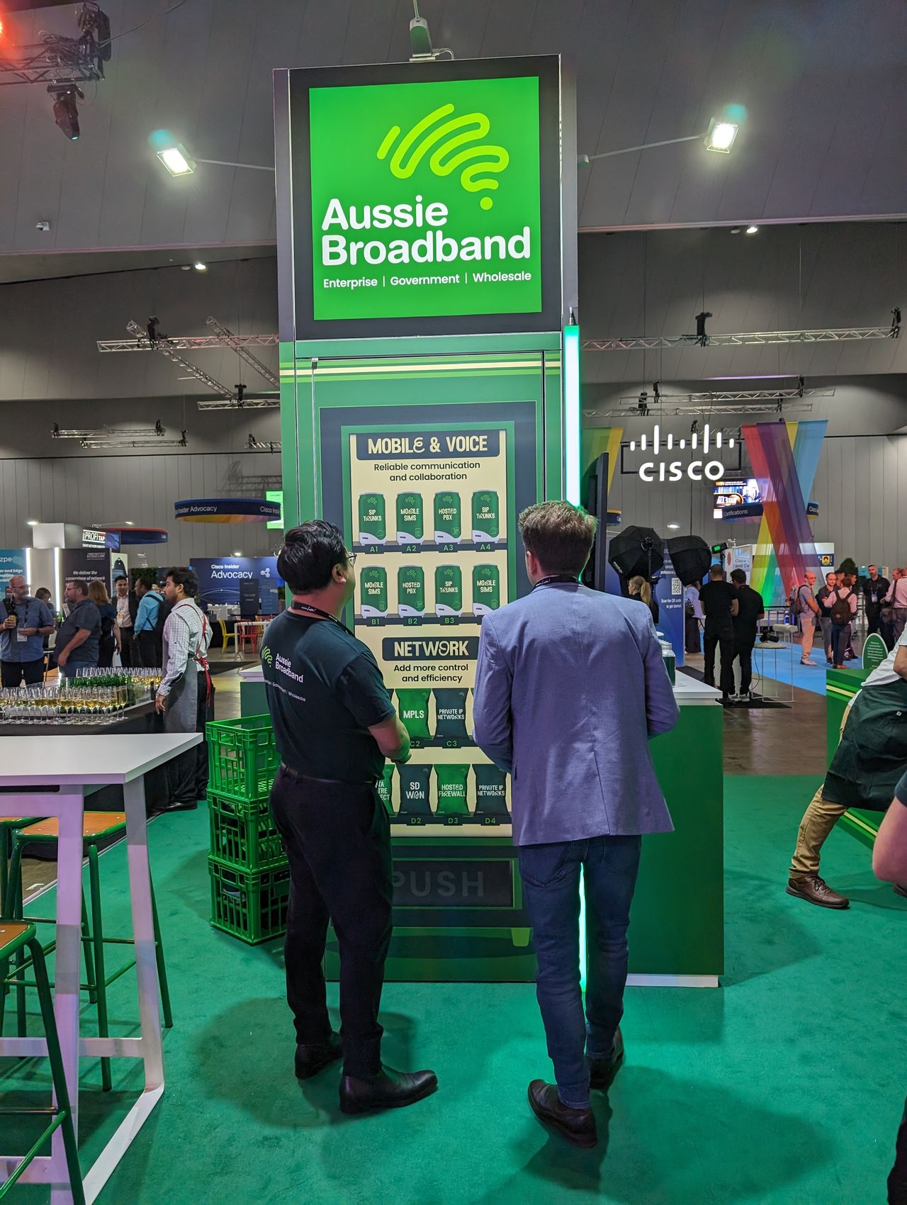 Aussie Broadband convenience store: Aussie Broadband's Cisco Live 2023 booth: A trade show booth resembling a convenience store. Two people, one wearing an Aussie Broadband shirt, arew staring at a sign meant to resemble a vending machine. Aussie Broadband Enterprise Technology offerings are listed on graphics of cans on the sign. "Network, add more control and efficiency", and "Mobile and Voice: reliable communication and collaboration".